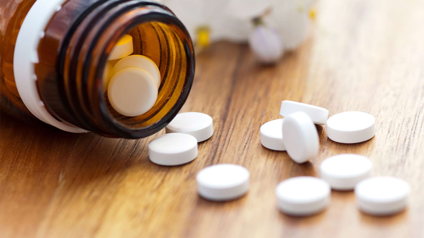 Schüßler-Salze – das steckt dahinter Schüßler-Salze: Braunes Gläschen mit Schüßler-Salz-Tabletten liegt auf einem Holztisch, davor einzelne weiße Tabletten.