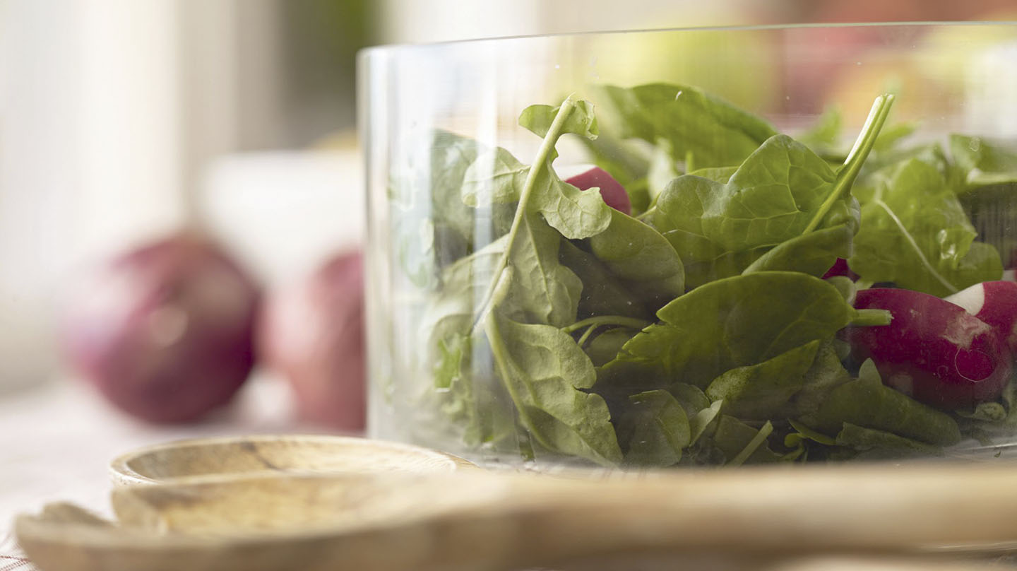 Mit den richtigen Lebensmitteln den Blutzucker im Lot Lebensmittel für konstanten Blutzucker: Glasschüssel mit Salatblättern und Radieschenstücken, davor liegt ein Salatbesteck aus Holz, im Hintergrund zwei rote Zwiebeln.