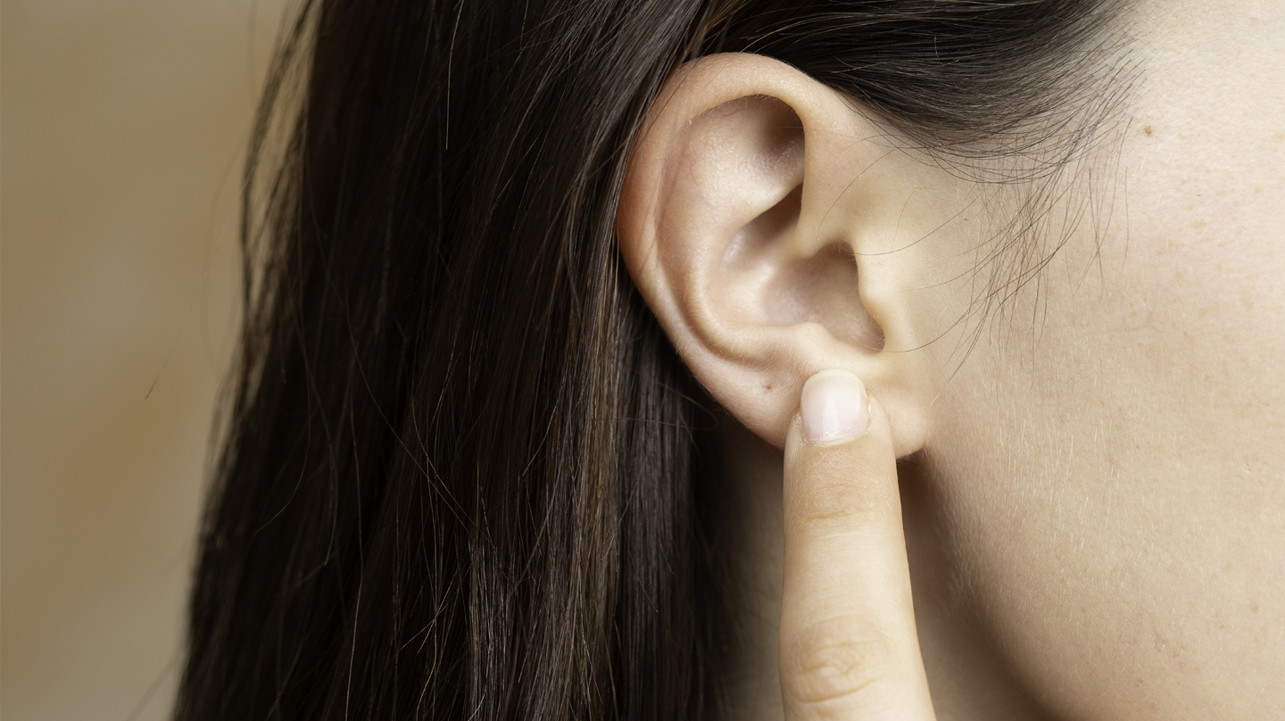 Ohren reinigen – so geht's ohne Stäbchen Ohren reinigen: Ein Zeigefinger zeigt auf das Ohr einer Frau mit braunen Haaren.